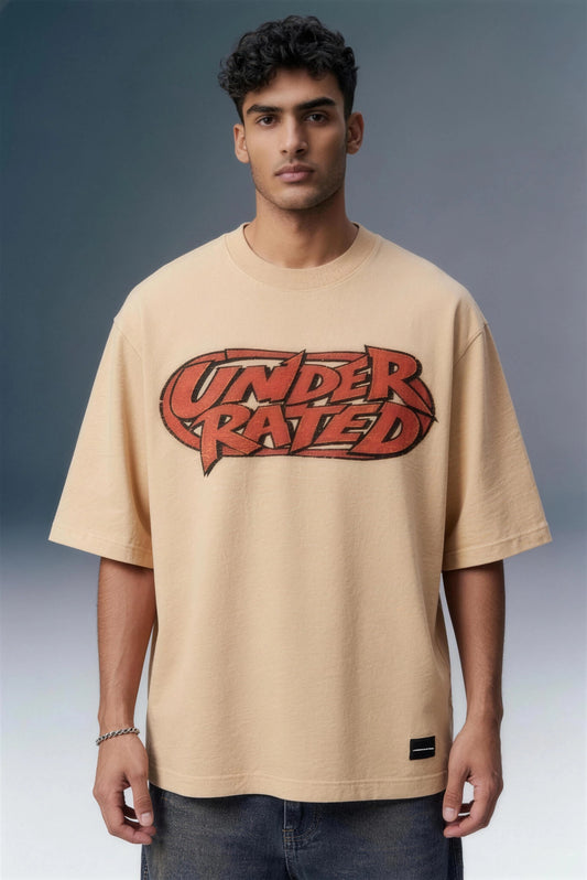 A man wearing a beige oversized t-shirt with a red and black 'UNDER RATED' graphic on the chest, paired with dark blue jeans, set against a gray studio background.