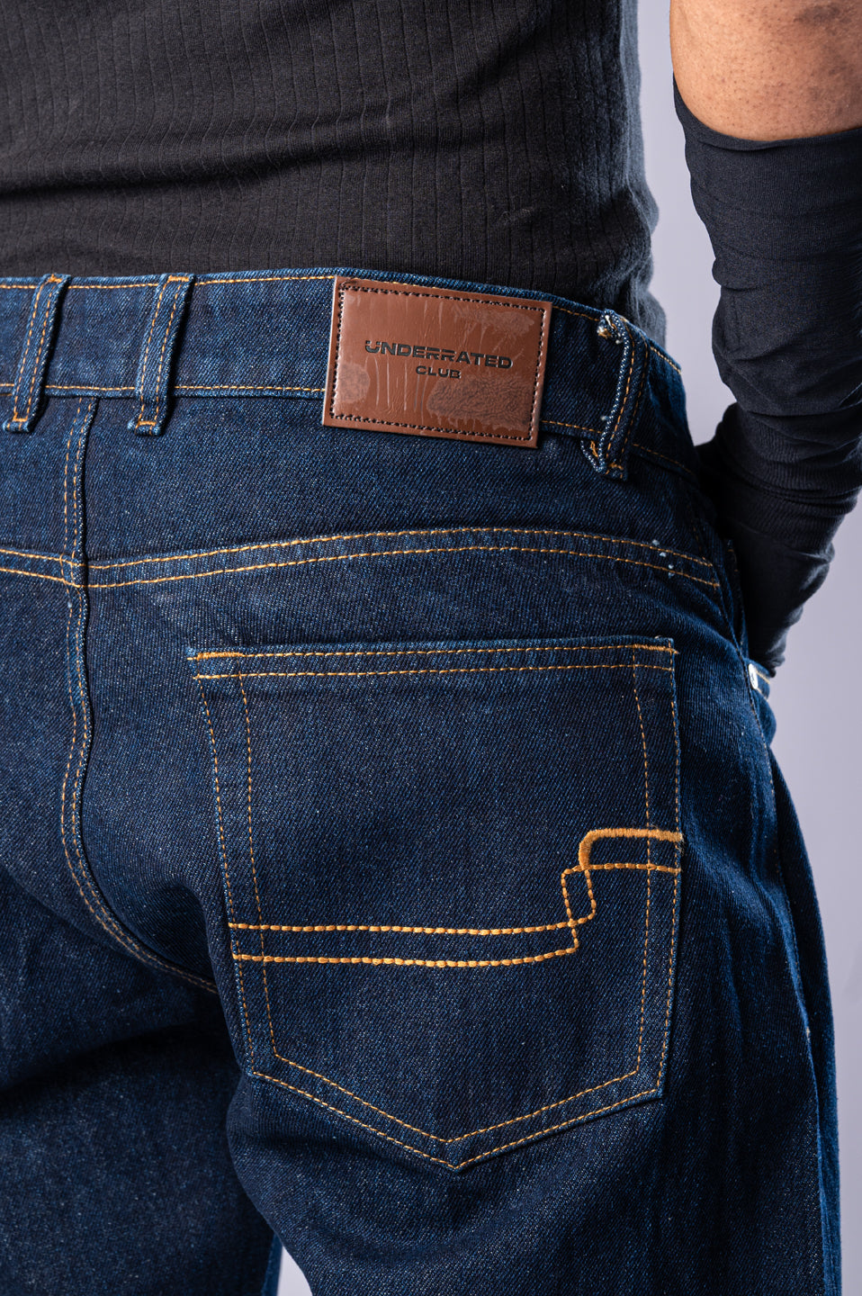 Close-up of the back of the dark-wash blue jeans, showing the brown leather Underrated Club patch on the waistband and detailed stitching on the back pocket.