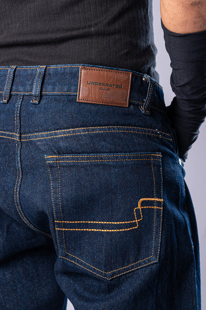 Close-up of the back of the dark-wash blue jeans, showing the brown leather Underrated Club patch on the waistband and detailed stitching on the back pocket.