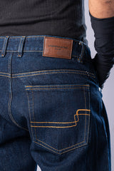 Close-up of the back of the dark-wash blue jeans, showing the brown leather Underrated Club patch on the waistband and detailed stitching on the back pocket.