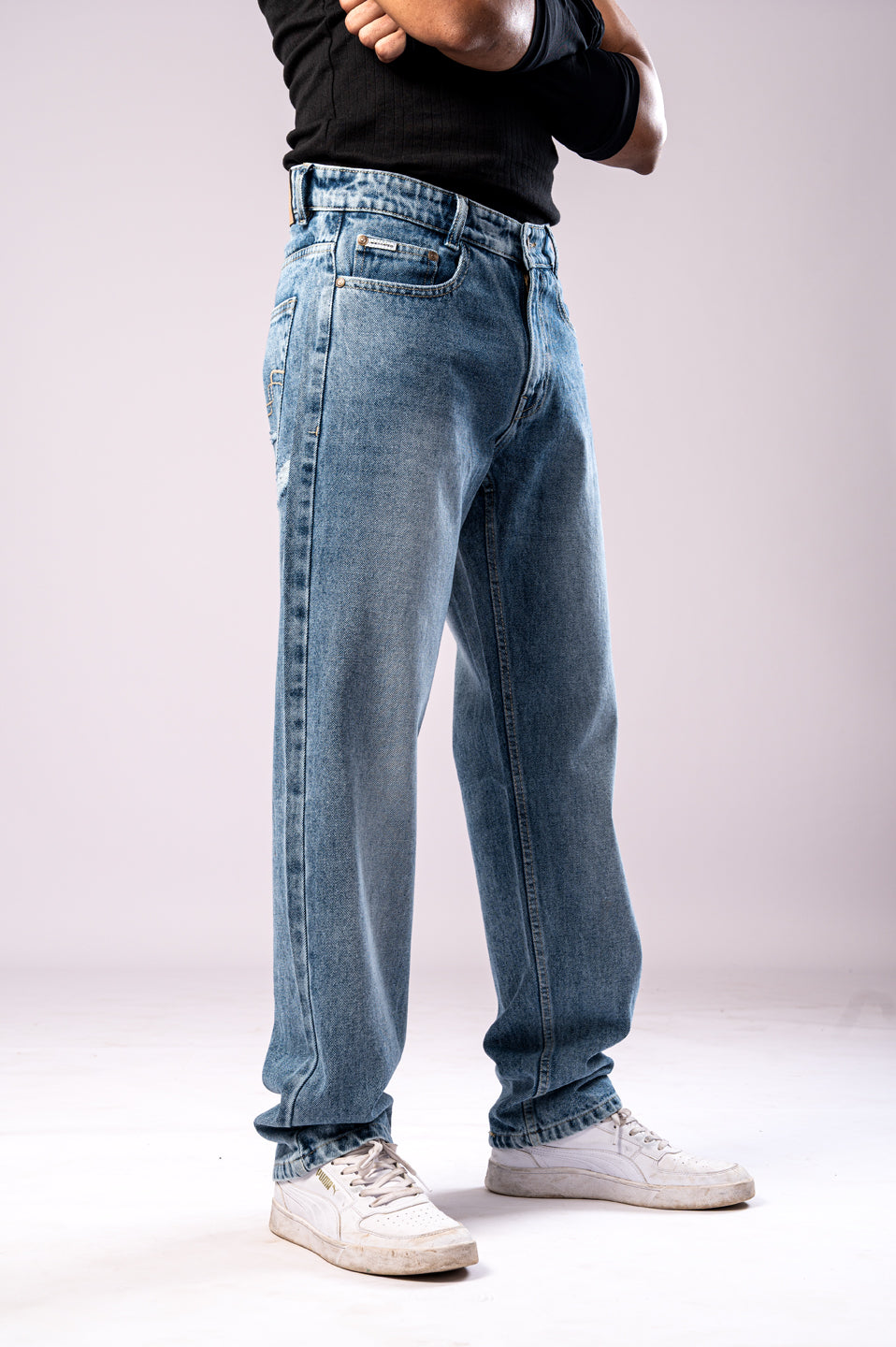 Angled side view of a person wearing light-wash blue baggy jeans, a black ribbed top, black arm sleeves, and white sneakers, with their arms crossed.