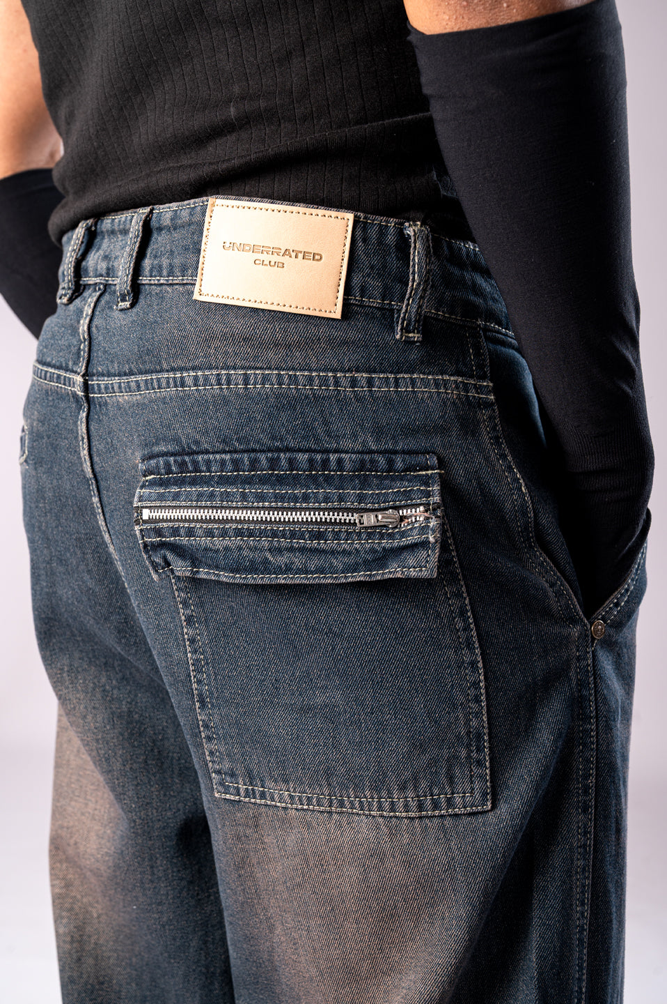 Close-up of the back of a person wearing dark, rusty-blue baggy cargo jeans, showing a back pocket with a horizontal zipper and a tan Underrated Club leather patch on the waistband.