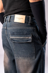 Close-up of the back of a person wearing dark, rusty-blue baggy cargo jeans, showing a back pocket with a horizontal zipper and a tan Underrated Club leather patch on the waistband.