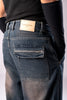 Close-up of the back of a person wearing dark, rusty-blue baggy cargo jeans, showing a back pocket with a horizontal zipper and a tan Underrated Club leather patch on the waistband.