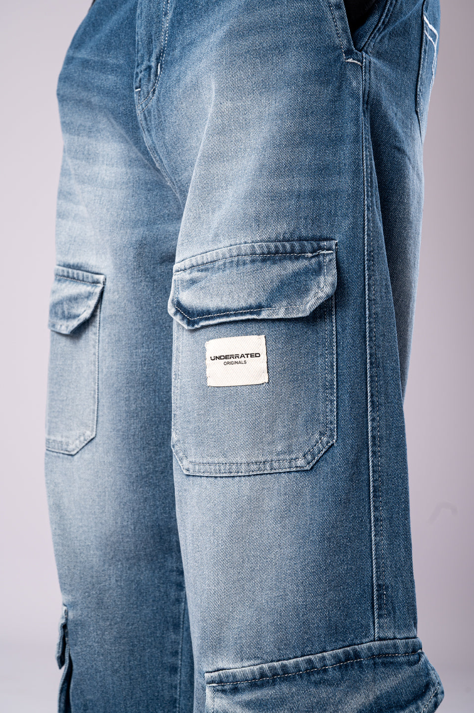 Close-up of the side cargo pocket on light-wash blue jeans, featuring a white Underrated Club logo patch.