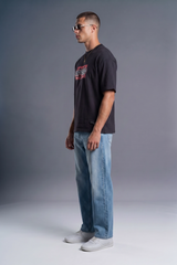 A man wearing a black graphic t-shirt, light blue jeans, and white sneakers, standing in profile against a gray studio background.