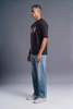 A man wearing a black graphic t-shirt, light blue jeans, and white sneakers, standing in profile against a gray studio background.