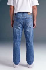 Rear view of a person wearing light blue denim jeans with a white t-shirt and white sneakers, standing against a gray studio background.