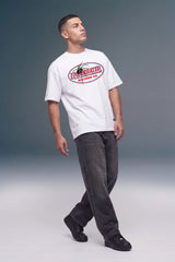A man wearing a white crewneck t-shirt with a red and black 'Unleashed Magazine Co' graphic on the chest, paired with dark gray jeans and black shoes, standing against a gray studio background.