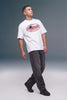A man wearing a white crewneck t-shirt with a red and black 'Unleashed Magazine Co' graphic on the chest, paired with dark gray jeans and black shoes, standing against a gray studio background.