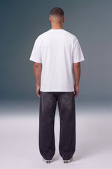 A man viewed from behind wearing a plain white short-sleeve t-shirt and dark charcoal gray baggy jeans, standing against a gray gradient studio background.