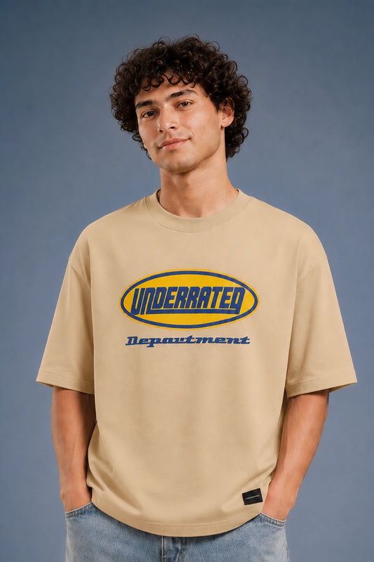 A man wearing a beige Under Rated Department t-shirt with a blue and yellow graphic logo on the chest, paired with blue jeans, standing against a blue-gray studio background.