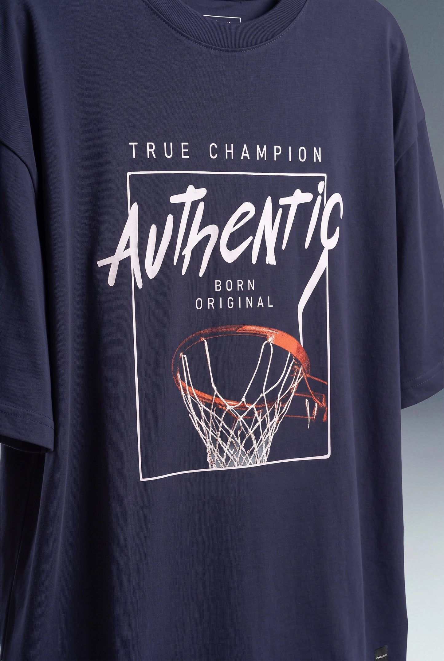 A navy blue oversized t-shirt featuring a basketball-themed graphic print with the text 'TRUE CHAMPION Authentic BORN ORIGINAL' and an orange basketball hoop illustration.