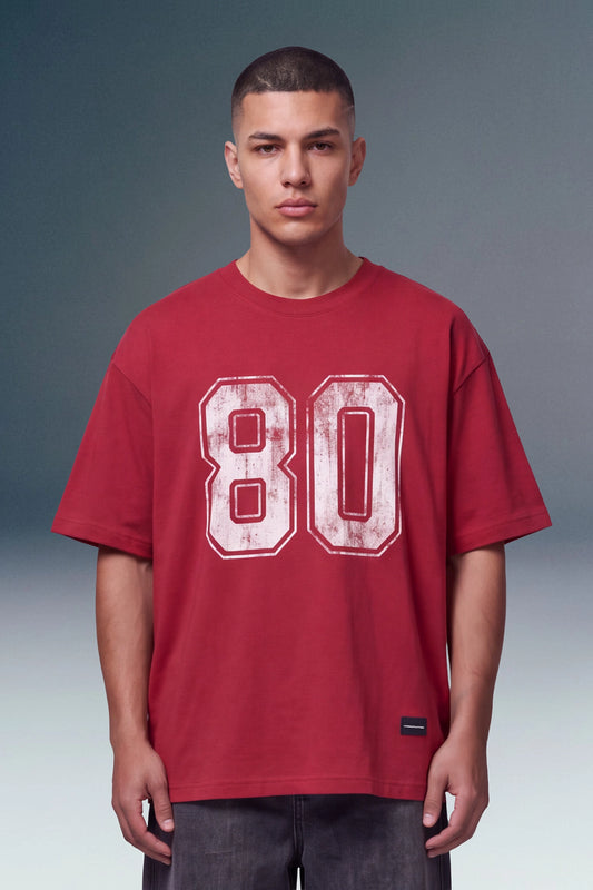 A man wearing a red oversized t-shirt with a distressed white '80' graphic on the front, photographed against a studio background with a gray-to-blue gradient.