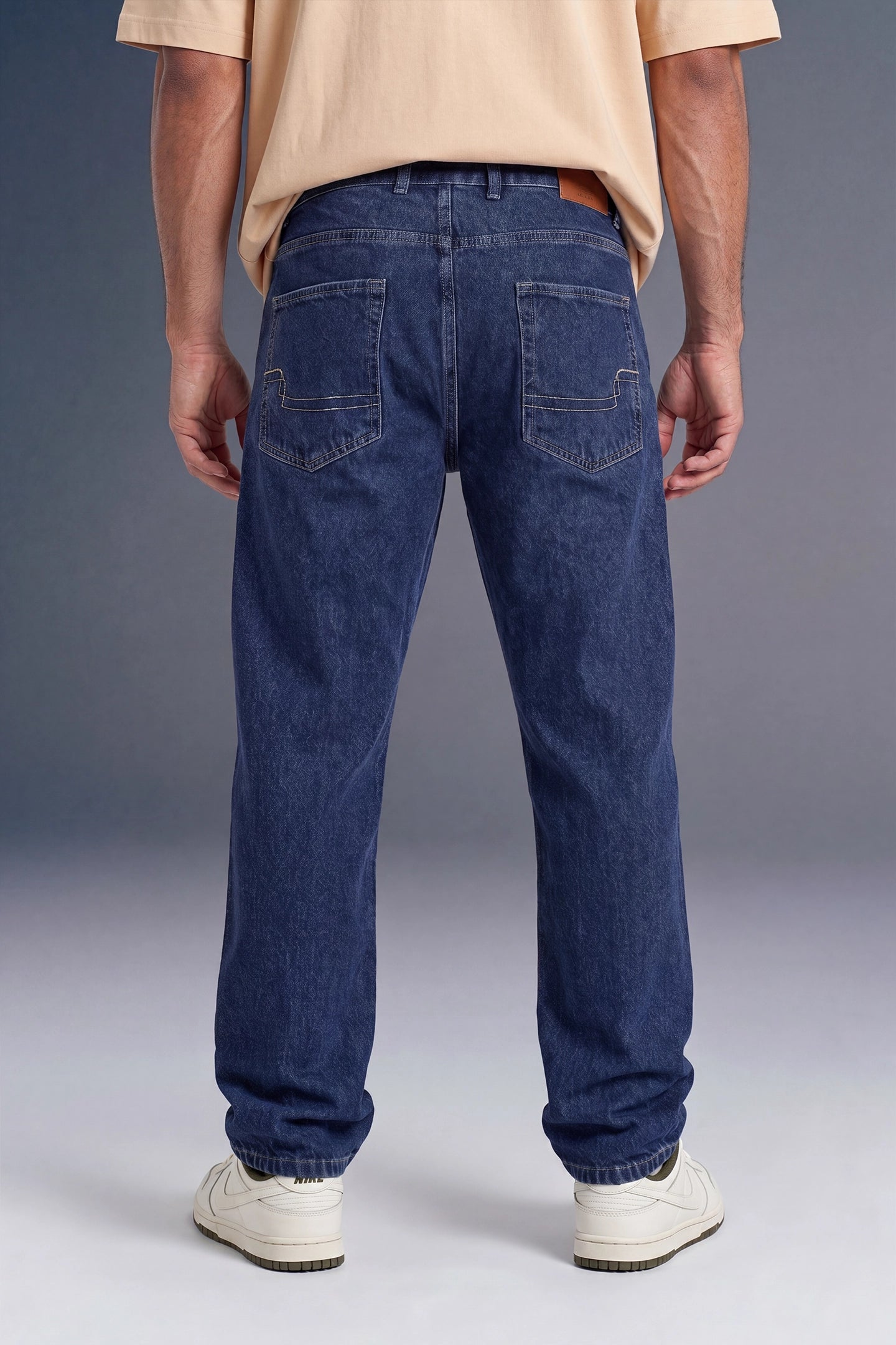 Rear view of a person wearing dark blue straight-leg jeans, a beige t-shirt, and white sneakers against a gray studio background.