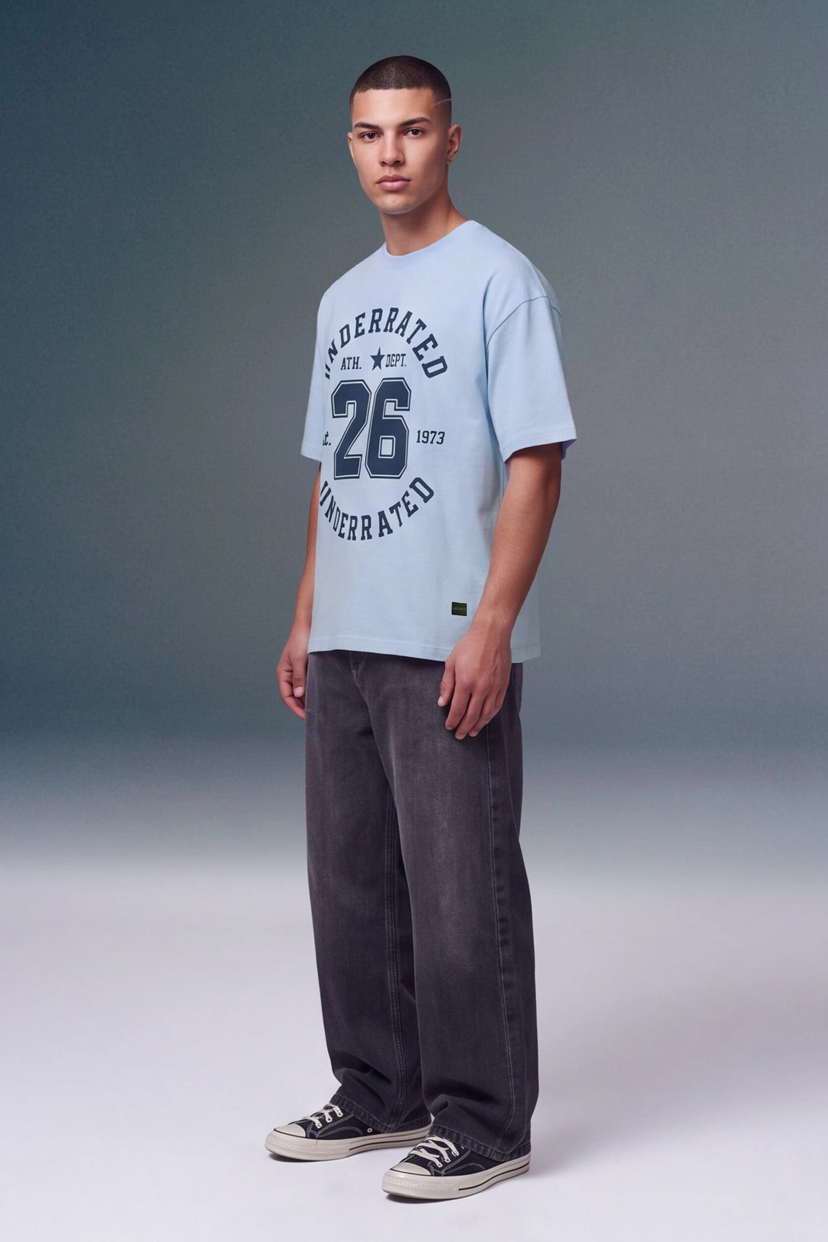 A male model wearing a light blue t-shirt with a dark navy graphic print that reads 'UNDERBATED' and '26', paired with dark gray jeans and black canvas sneakers, standing against a gray studio background.