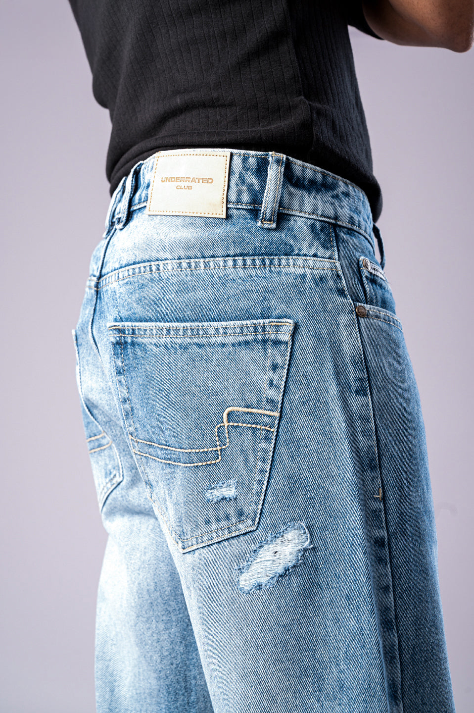 Close-up of the back of a person wearing light-wash blue ripped jeans, showing a distressed patch on the leg and a tan Underrated Club leather patch on the waistband.
