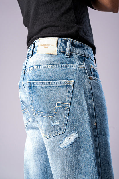 Close-up of the back of a person wearing light-wash blue ripped jeans, showing a distressed patch on the leg and a tan Underrated Club leather patch on the waistband.
