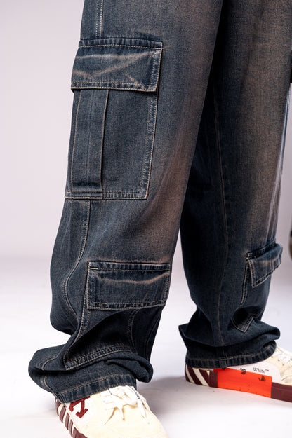 Close-up side view of the leg of a person wearing dark, rusty-blue baggy cargo jeans, showing the detail and texture of the stacked cargo pockets.