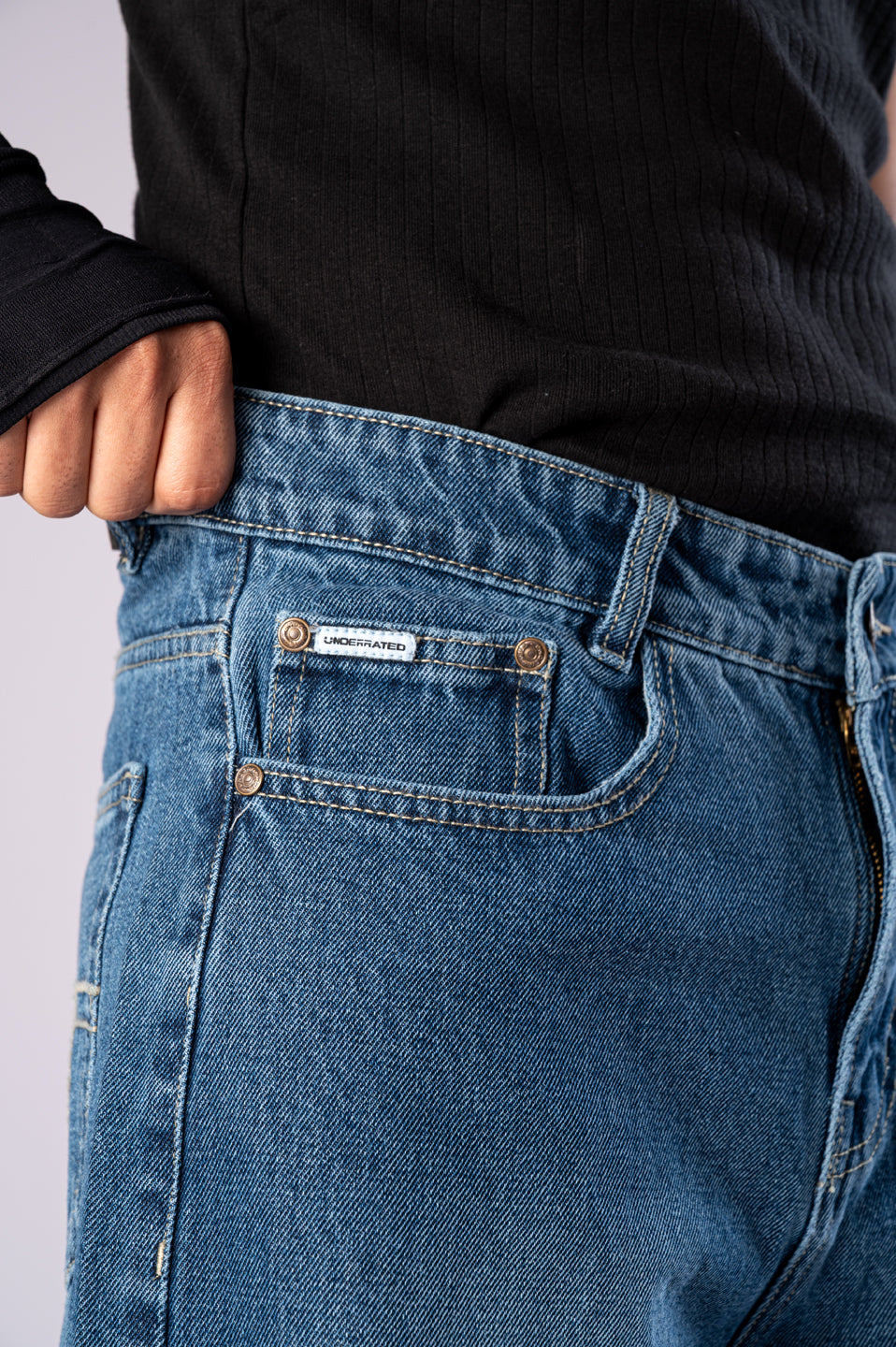 Close-up of the front pocket of dark-wash blue jeans, showing a small white "UNDERRATED CLUB" logo tag.