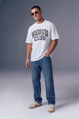 A man wearing a white graphic t-shirt with 'UNDERRATED CLUB' printed in navy and gold, paired with blue jeans and beige sneakers, standing against a gray studio background.