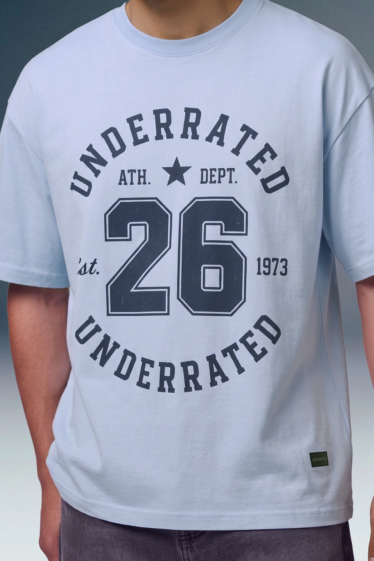 A close-up of a person wearing a light blue t-shirt with a dark navy graphic print on the front that reads 'UNDERRATED ATH. DEPT.