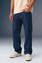 A person wearing dark blue straight-leg jeans, a light beige t-shirt, and off-white sneakers, shown from the waist down against a studio background.