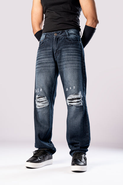 Angled side view of a person wearing dark-wash blue baggy jeans with large, distressed rips on both knees, a black top, and black sneakers.