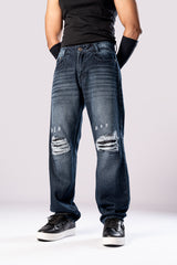 Angled side view of a person wearing dark-wash blue baggy jeans with large, distressed rips on both knees, a black top, and black sneakers.