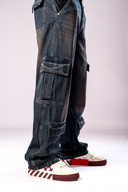 Side view of a person's legs wearing dark, rusty-blue baggy cargo jeans with multiple stacked cargo pockets and white and maroon sneakers.