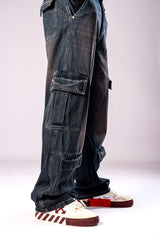 Side view of a person's legs wearing dark, rusty-blue baggy cargo jeans with multiple stacked cargo pockets and white and maroon sneakers.