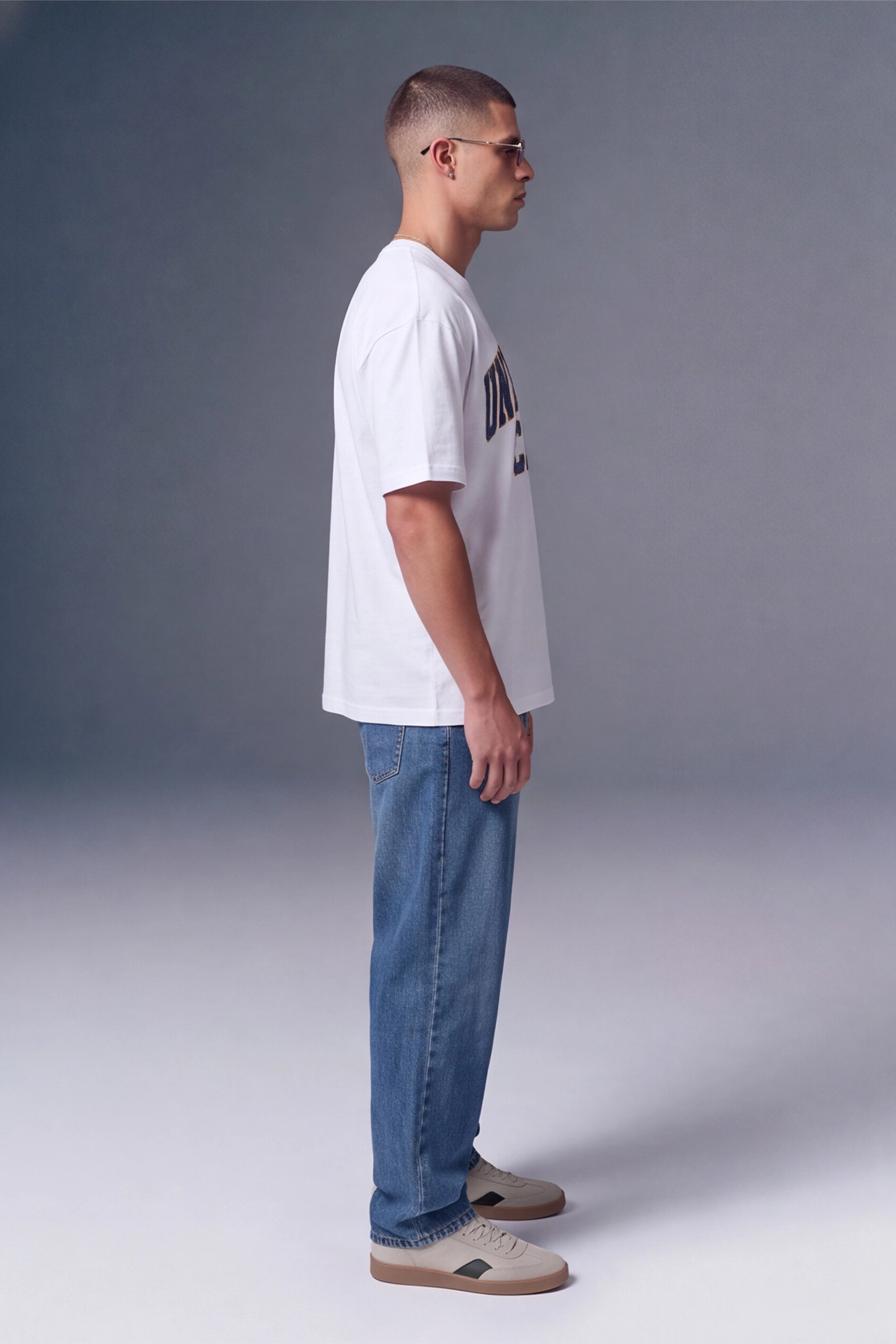 A man in profile wearing a white graphic t-shirt, blue jeans, and beige sneakers, standing against a gray studio background.