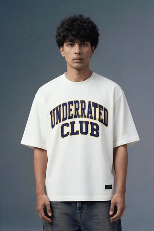 A man wearing a white crewneck t-shirt with 'UNDERRATED CLUB' printed in navy blue varsity lettering on the chest, paired with dark gray jeans, set against a dark gray studio background.