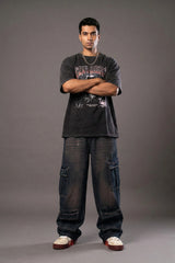 Man in a black graphic tee and baggy dark blue cargo jeans standing with arms crossed against a grey background.