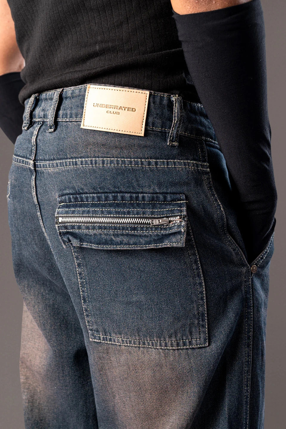 Close-up of the back pocket and waistband of blue denim jeans, showing a white "UNDERRATED" brand patch.