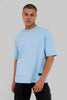 A man wearing a light blue oversized crewneck t-shirt and black cargo pants, standing against a solid gray studio background.
