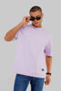 A man wearing a light purple short-sleeve crewneck t-shirt and blue jeans, posing with black sunglasses against a solid gray background.