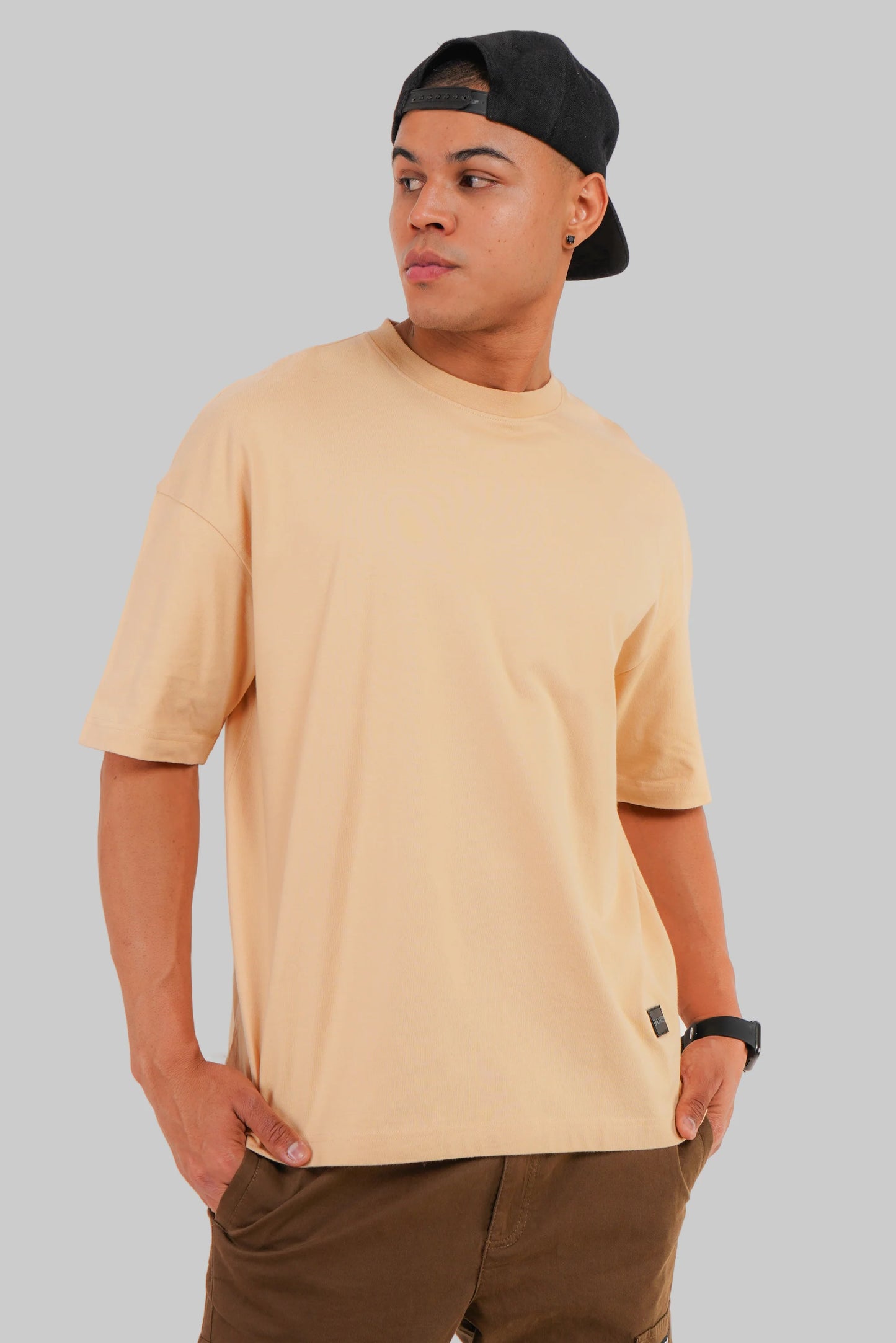 A man wearing a light beige crew-neck t-shirt, brown pants, and a black baseball cap, photographed against a solid gray background.