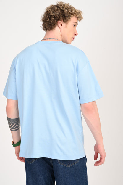 Back view of a young man with curly blonde hair wearing a light blue oversized t-shirt and dark wash wide-leg jeans. A silver chain necklace is visible, and he has a geometric tattoo on his left forearm.