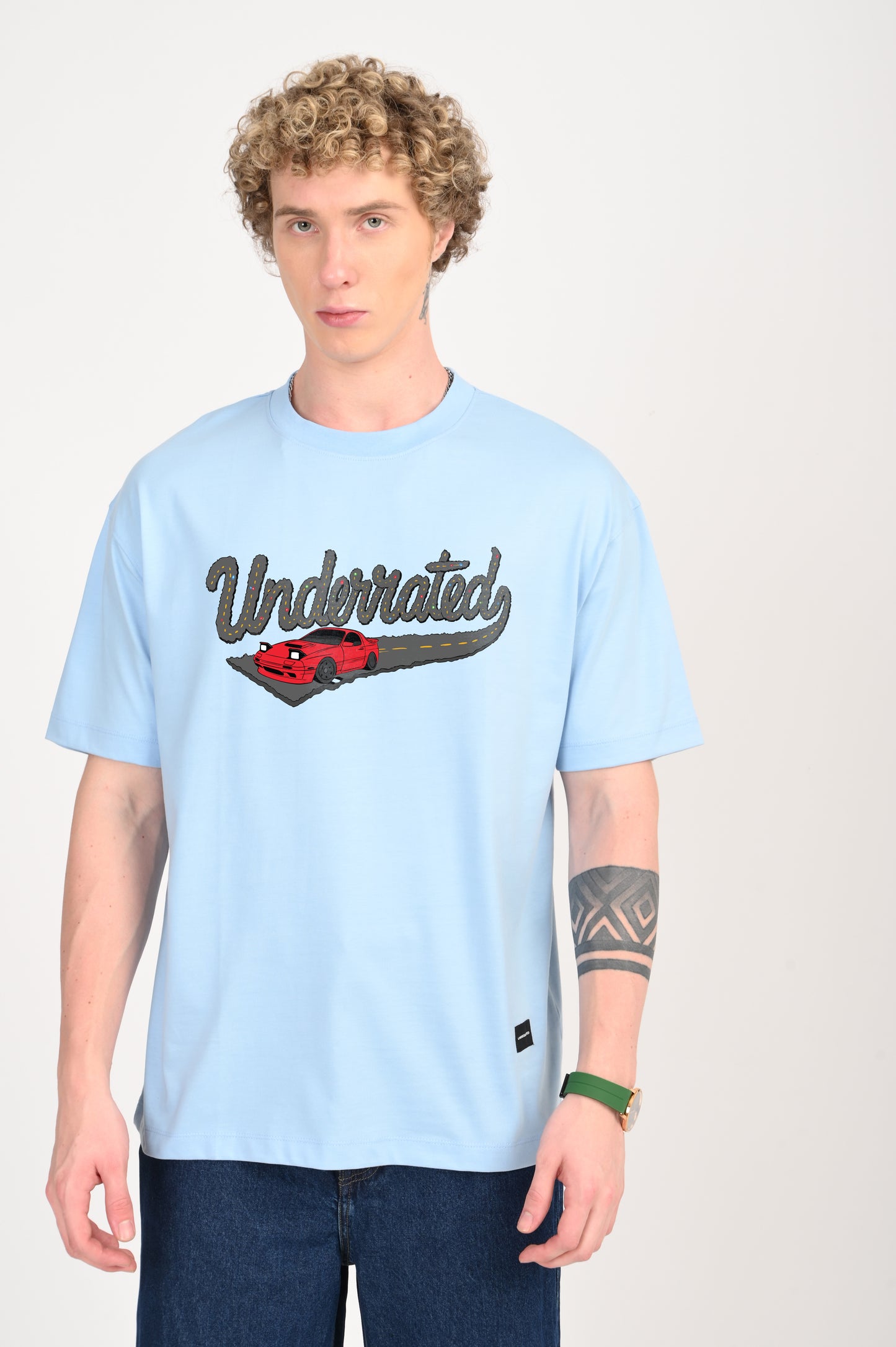 A full-frame portrait of a young man with curly blonde hair wearing a light blue oversized t-shirt featuring a graphic of a red car and the word "Underrated," paired with dark wash wide-leg jeans. He has a tattoo on his forearm and is wearing a gold watch with a green strap.