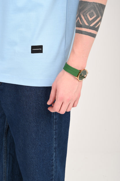 A close-up shot of a man's arm and torso, showing a light blue textured t-shirt, dark wash denim jeans, a gold watch with a green strap, and a geometric tattoo on his forearm.