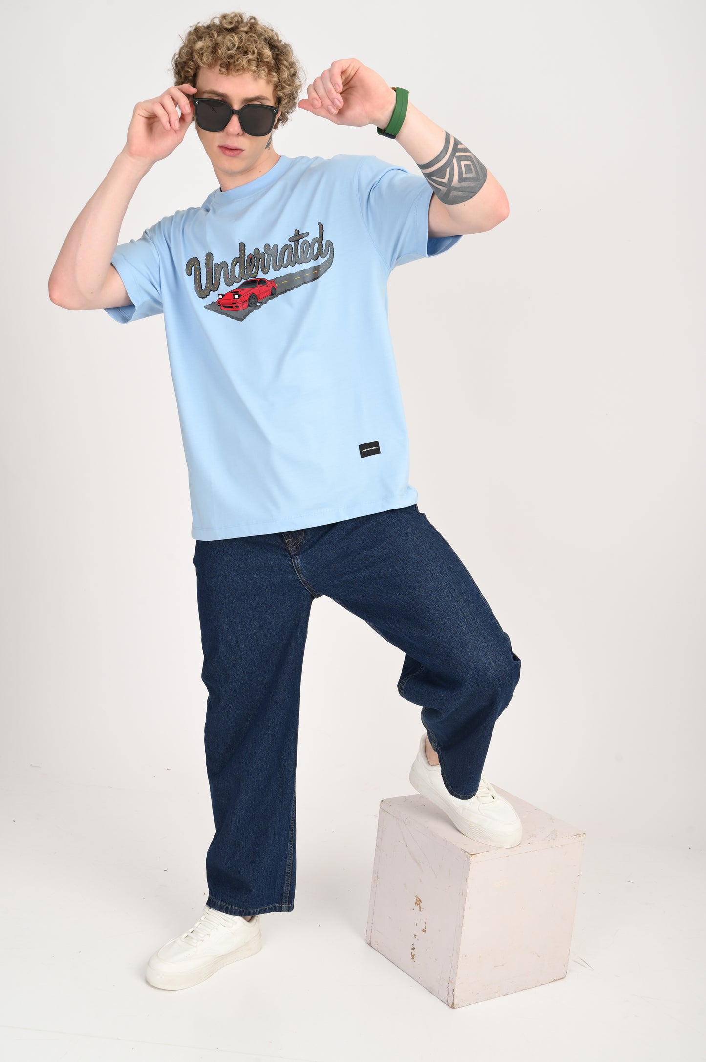 A young man with curly blonde hair wearing a light blue oversized t-shirt with a graphic of a red car and the word "Underrated," dark wash wide-leg jeans, and white sneakers. He is adjusting his black square-frame sunglasses and standing with one foot on a white block.