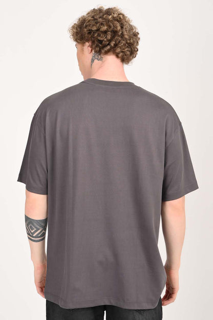 Back view of a young man with curly blonde hair wearing a plain charcoal gray oversized t-shirt and dark trousers. He has a geometric tattoo on his left forearm and a neck tattoo.
