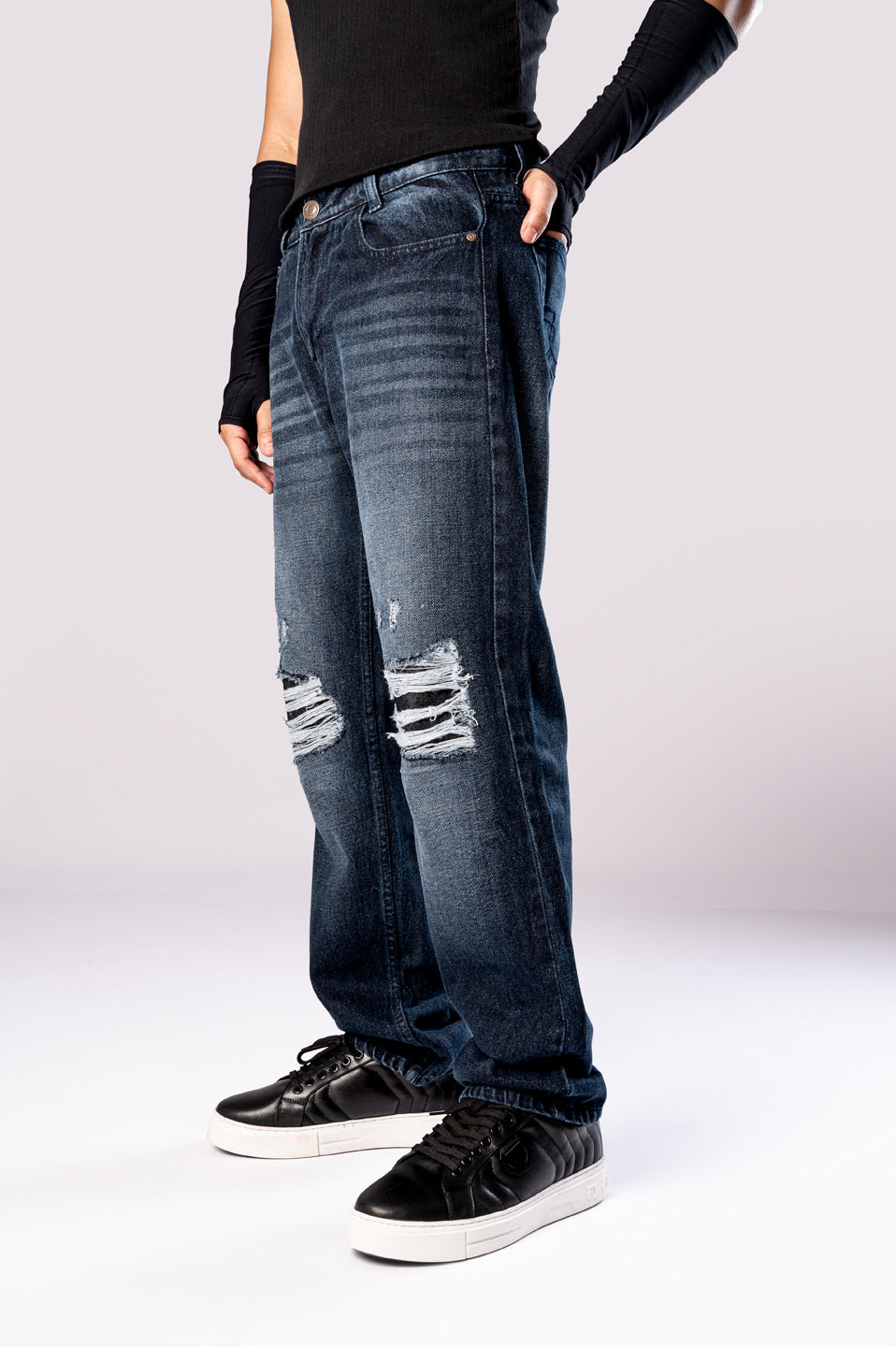 Angled side view of a person wearing dark-wash blue baggy jeans with ripped knees, a black top, and black sneakers.