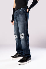 Angled side view of a person wearing dark-wash blue baggy jeans with ripped knees, a black top, and black sneakers.