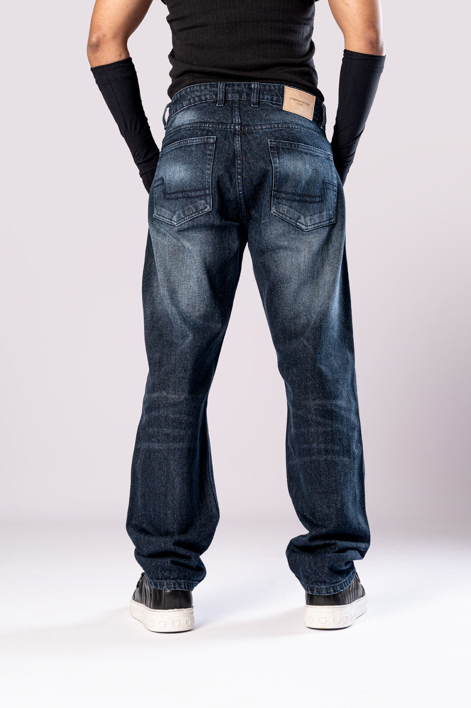 Full-body front view of a person wearing dark-wash blue baggy jeans with ripped knees, a black top, and black arm sleeves, with their hands in their pockets.