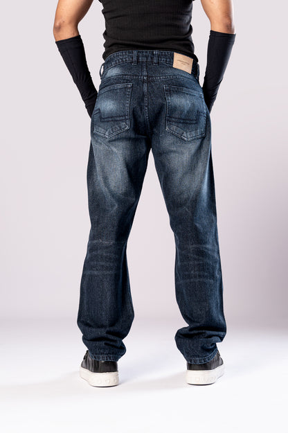 Full-body front view of a person wearing dark-wash blue baggy jeans with ripped knees, a black top, and black arm sleeves, with their hands in their pockets.