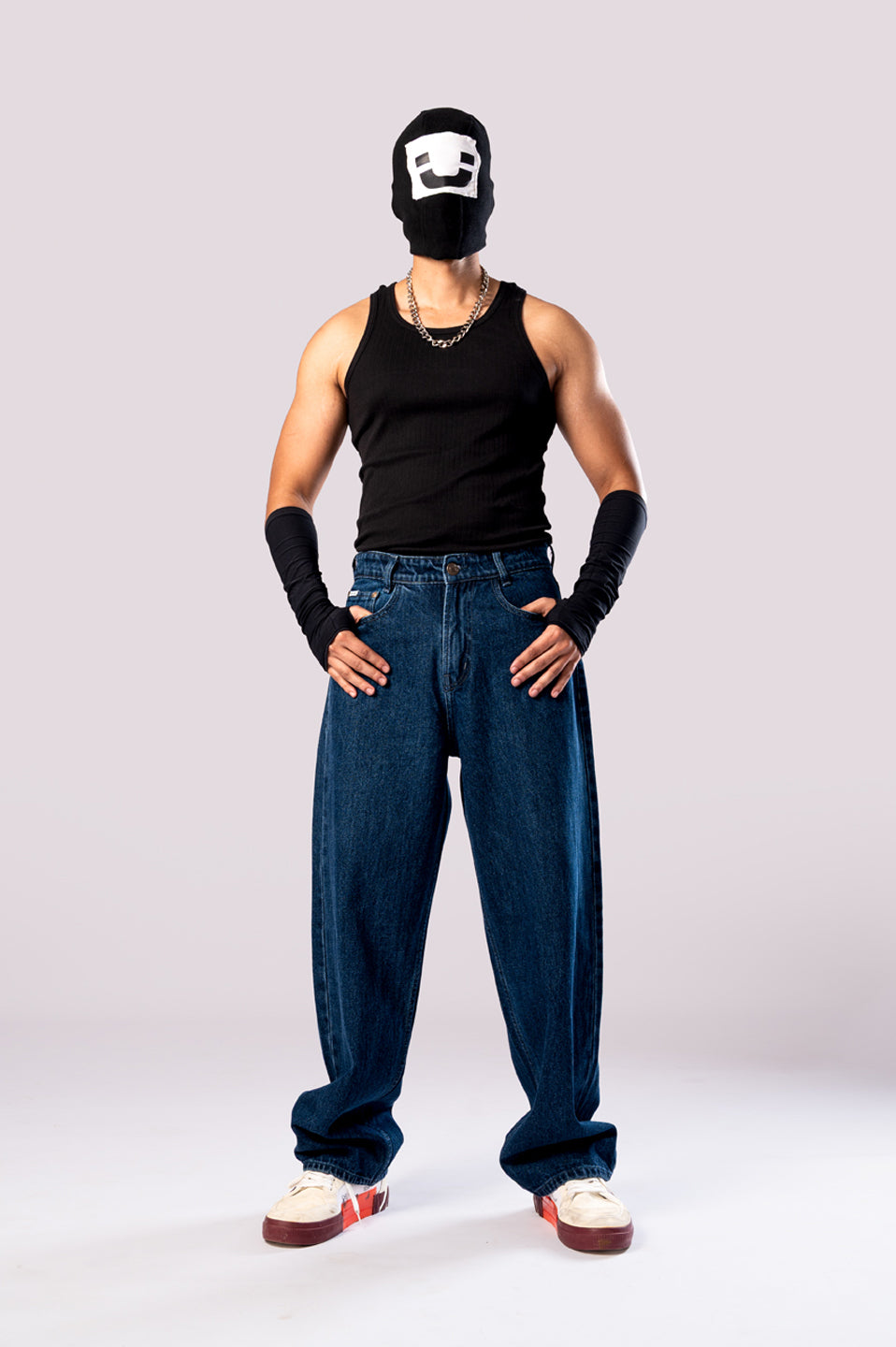 Full-body front view of a person wearing baggy, dark blue jeans with a black tank top, black arm sleeves, and a white cap.