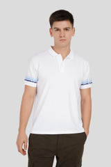 A man wearing a white polo shirt with blue striped cuffs and dark trousers against a neutral grey background.