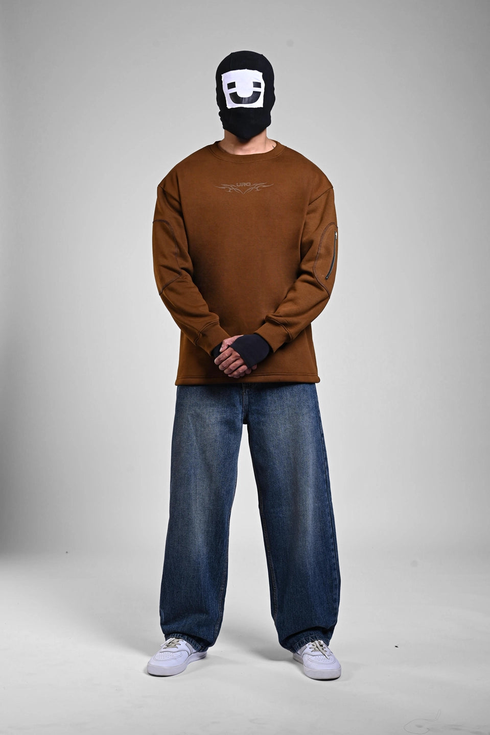 Front view of a person in a brown sweatshirt, baggy blue jeans, and a white-on-black graphic balaclava.